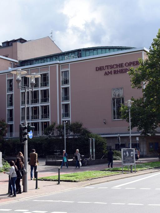 Das Opernhaus in Düsseldorf- ein großes ockerfarbenes Gebäude. Daneben ist eine Straße. Im Vordergund sind Passanten zu sehen. 