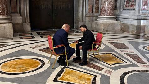 Das Foto zeigt US-Präsident Trump und den ukrainischen Präsidenten Selenskyj im Gespräch miteinander. Beide sitzen sich gegenüber auf goldenen Stühlen, die mit rotem Stoff bezogen sind.