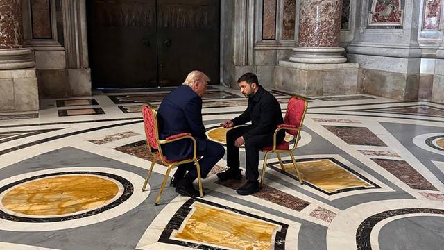 Das Foto zeigt US-Präsident Trump und den ukrainischen Präsidenten Selenskyj im Gespräch miteinander. Beide sitzen sich gegenüber auf goldenen Stühlen, die mit rotem Stoff bezogen sind.