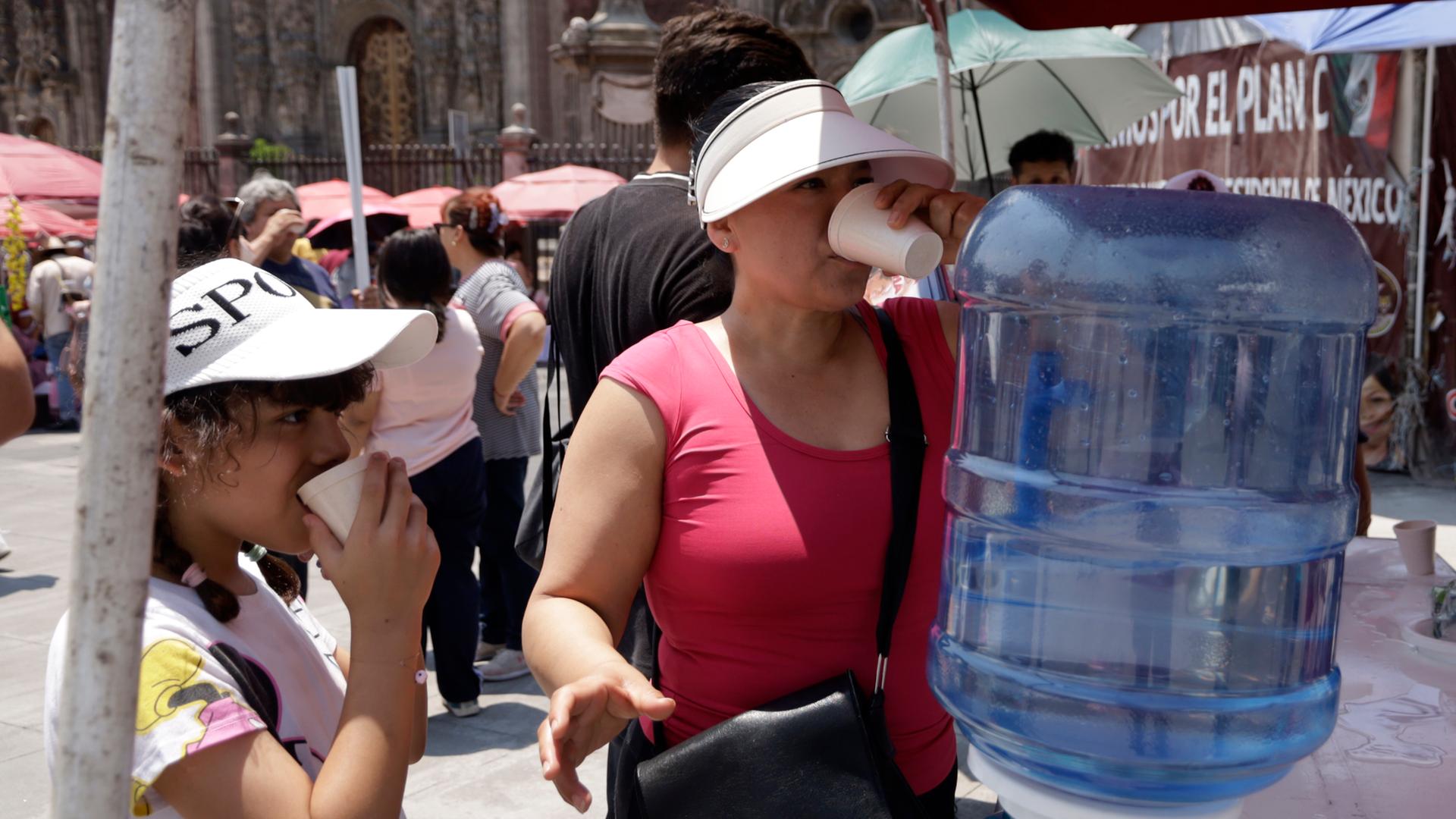 Zwei Frauen trinken Wasser vor einem Wasserspender. Im Hintergrund sind Menschen mit Sonnenschirmen zu sehen. Zwei Frauen trinken Wasser vor einem Wasserspender. Im Hintergrund sind Menschen mit Sonnenschirmen zu sehen.