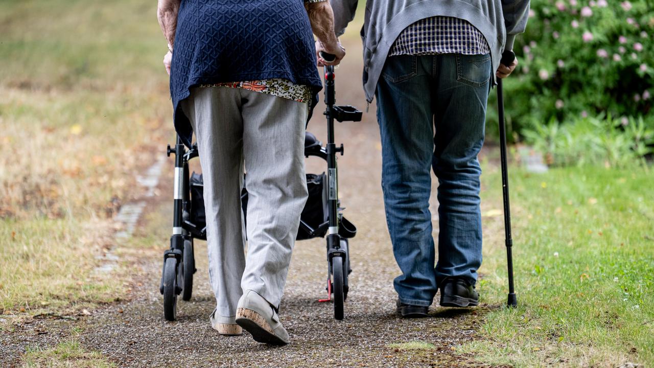 Zwei Senioren gehen mit Gehhilfen durch den Garten eines Seniorenheims. Eine Person nutzt einen Rollator, die andere einen Gehstock. 