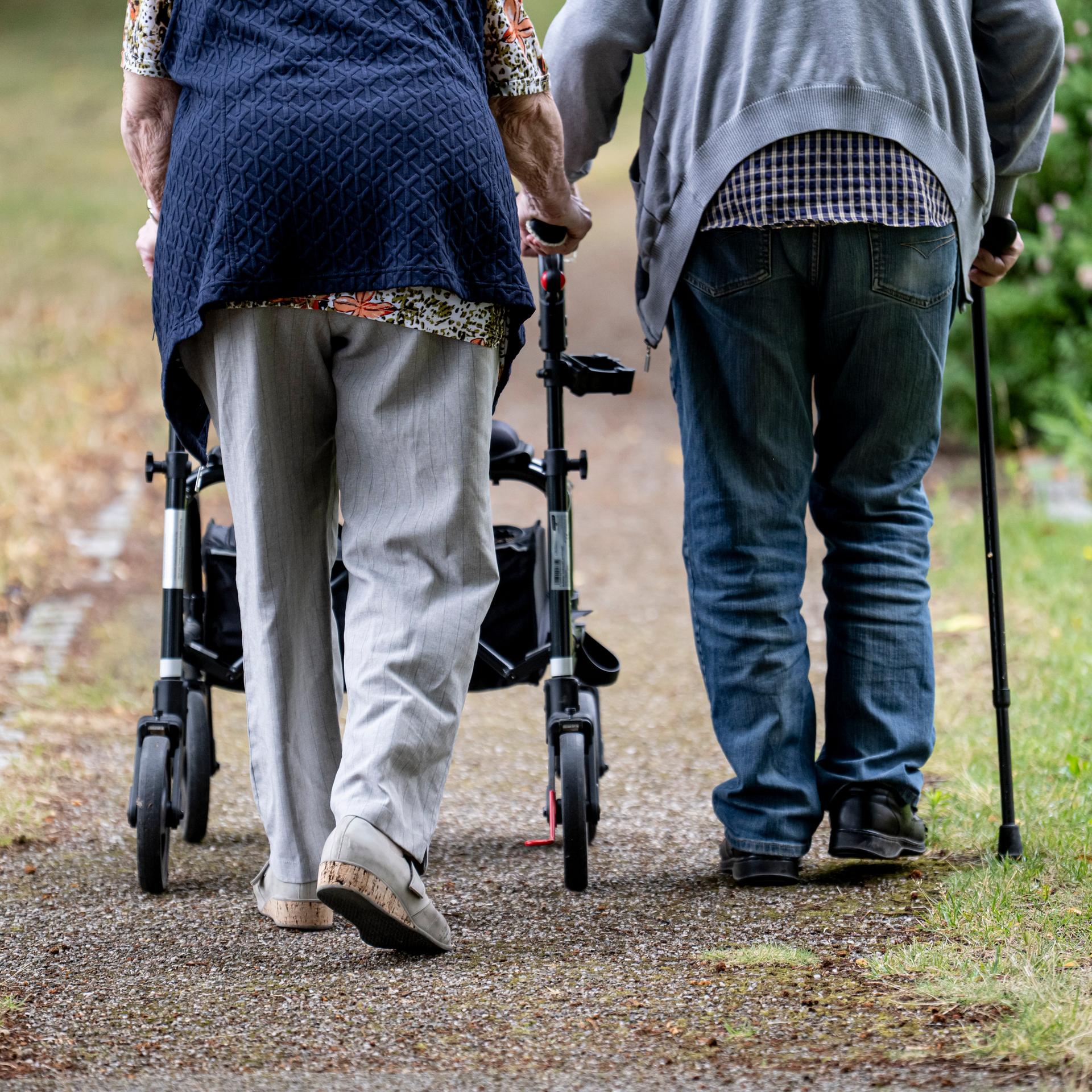 Zwei Senioren gehen mit Gehhilfen durch den Garten eines Seniorenheims. Eine Person nutzt einen Rollator, die andere einen Gehstock. 