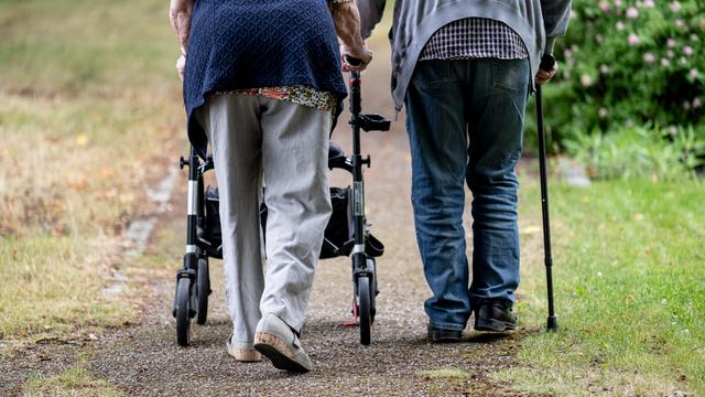 Zwei Senioren gehen mit Geh-Hilfen durch den Garten eines Senioren-Heims. Eine Person nutzt einen Rollator, die andere einen Geh-Stock. 