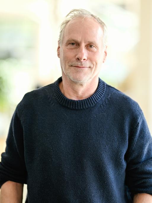 Der Schauspieler Wolfram Koch steht vor einer Pressekonferenz zum neuen Stück der Nibelungen-Festspiele. Er trägt einen dunkelblauen Pullover und schaut freundlich in die Kamera.