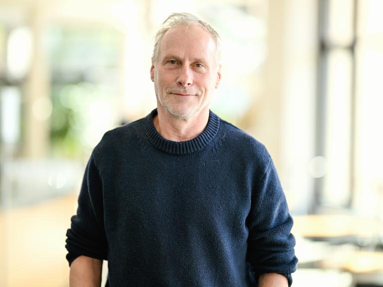 Der Schauspieler Wolfram Koch steht vor einer Pressekonferenz zum neuen Stück der Nibelungen-Festspiele. Er trägt einen dunkelblauen Pullover und schaut freundlich in die Kamera.