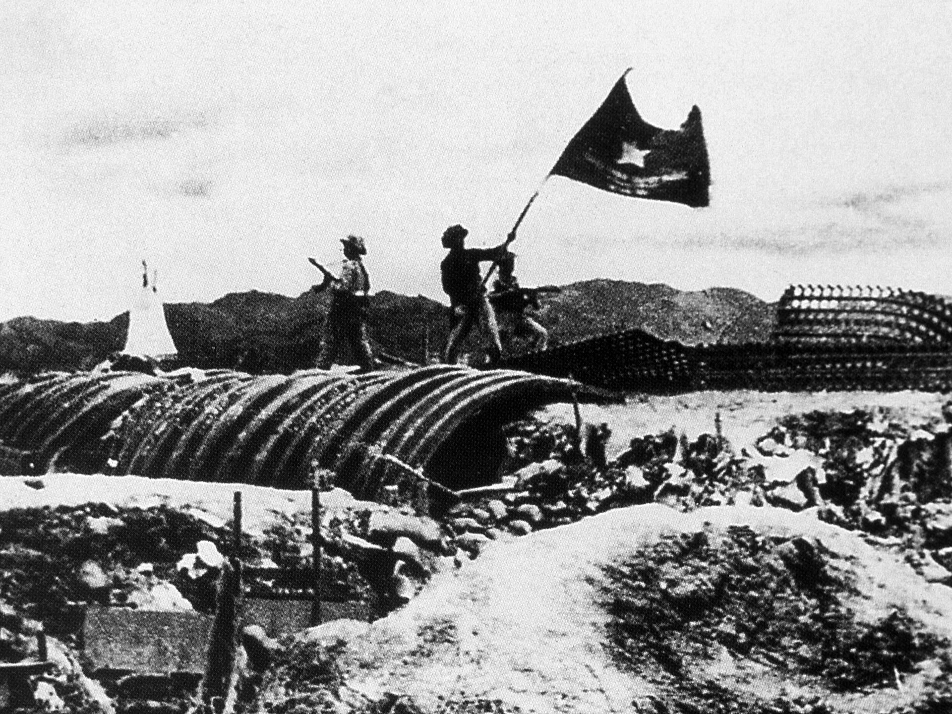 Eine historische Schwarzweiß-Aufnahme zeigt das Schlachtfeld von Dien Bien Phu in Vietnam. Ein Soldat hält die vietnamesische Flagge hoch.