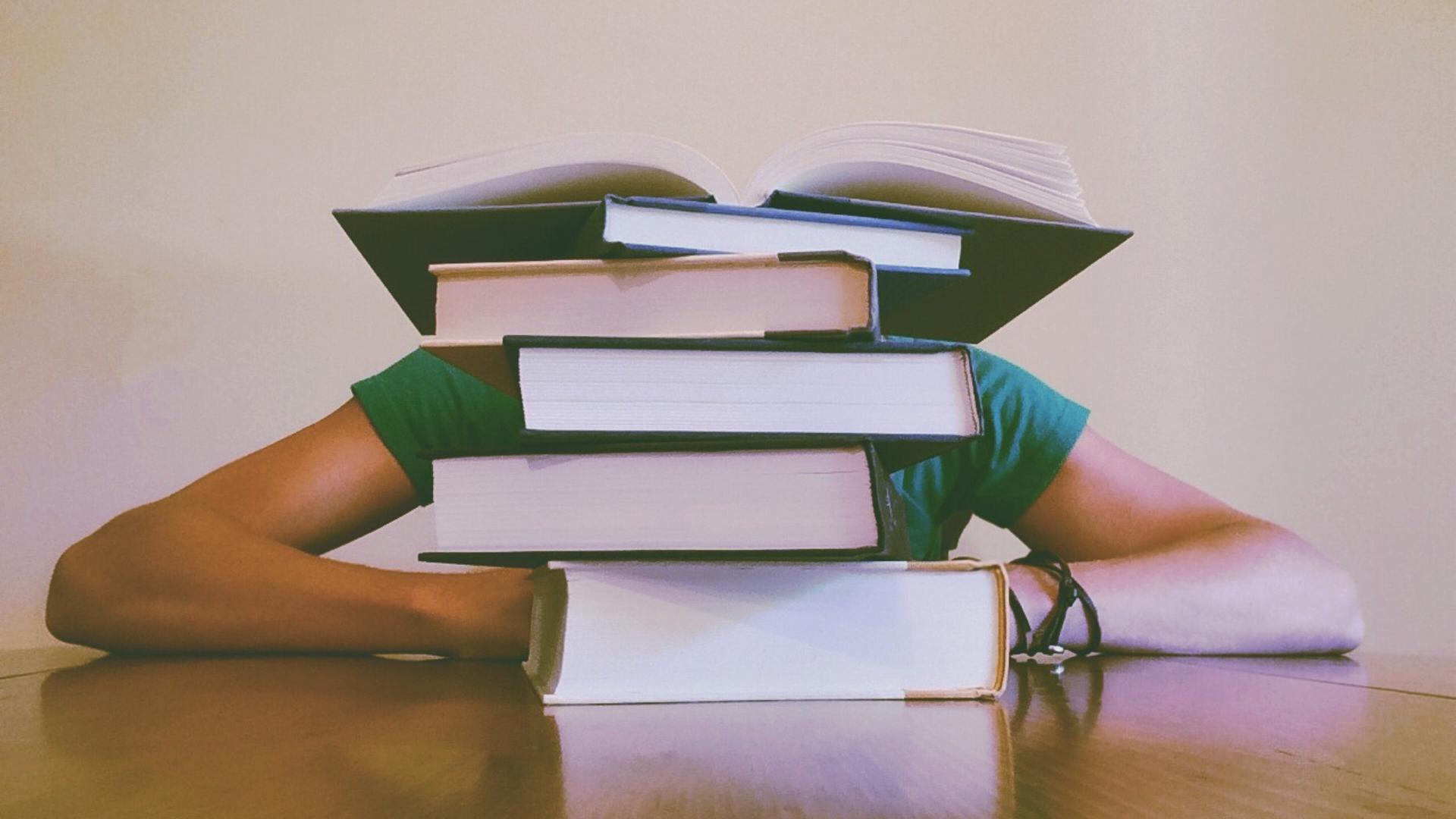 Ein Stapel Bücher liegt auf dem Tisch. Dahinter sitzt eine Person. Ein Stapel Bücher liegt auf dem Tisch. Dahinter sitzt eine Person.