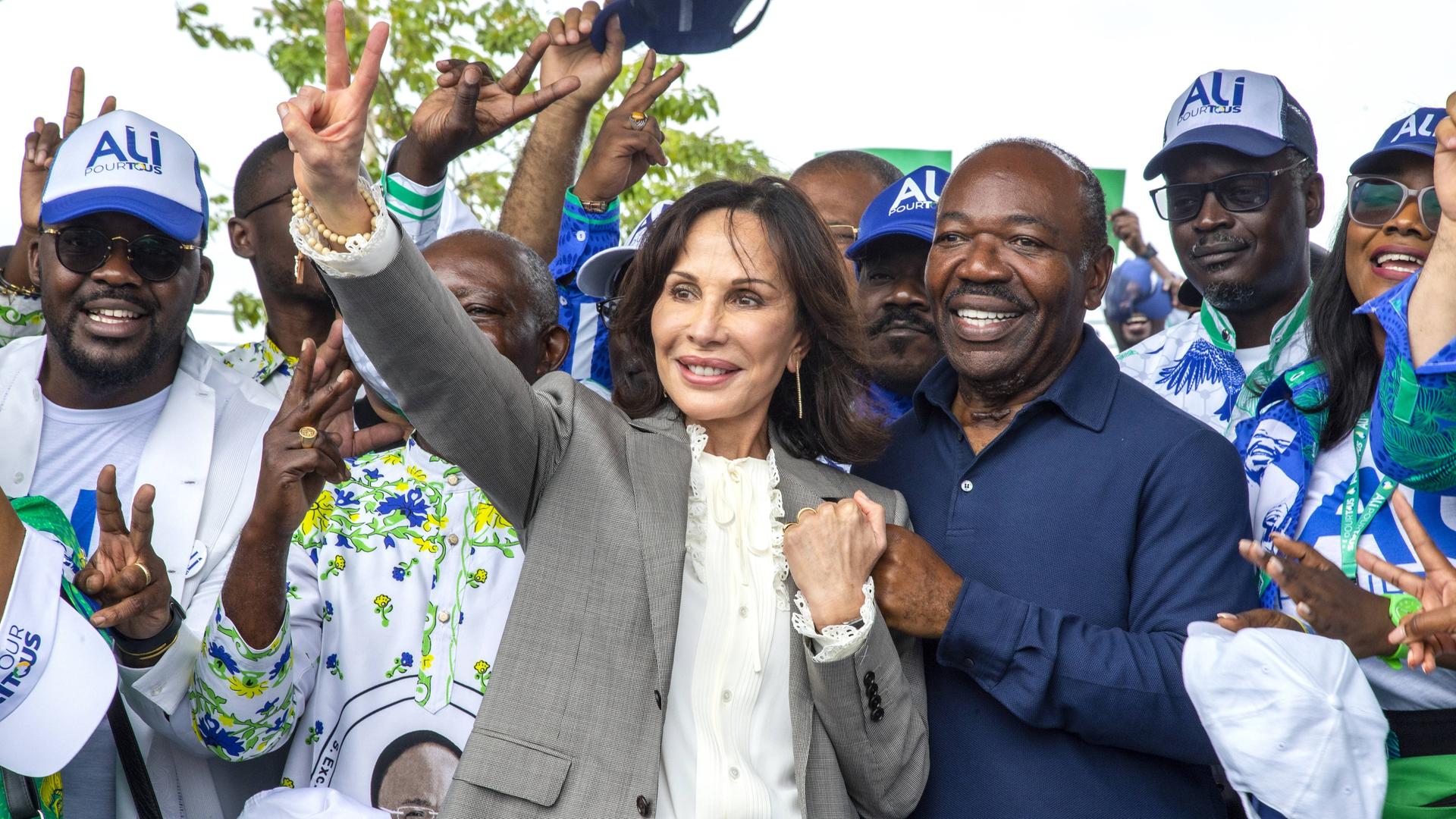 Sylvia Bongo winkt und lächertl, hinter ihr Ali Bongo. Beide sind von Anhängern umgeben. 