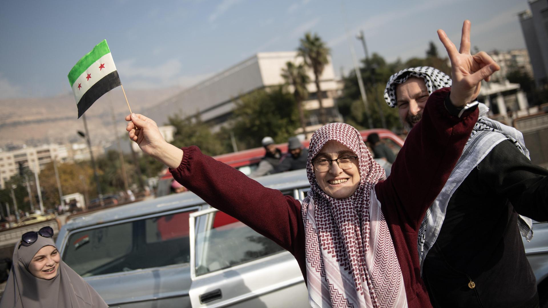 Menschen feiern auf den Straßen von Damaskus den Sturz von Machthaber Assad, eine Frau hält in einer Hand eine syrische Flagge, mit der anderen formt sie ein Victory-Zeichen.