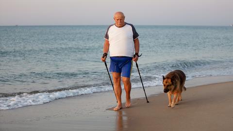 Symbolbild: Ein älterer Mann geht mit Stöcken und einem Hund am Meer entlang.