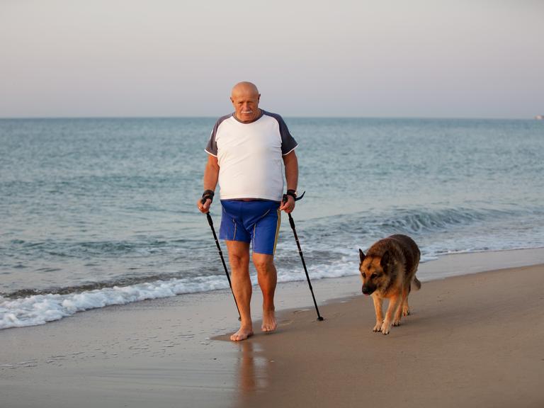 Symbolbild: Ein älterer Mann geht mit Stöcken und einem Hund am Meer entlang.