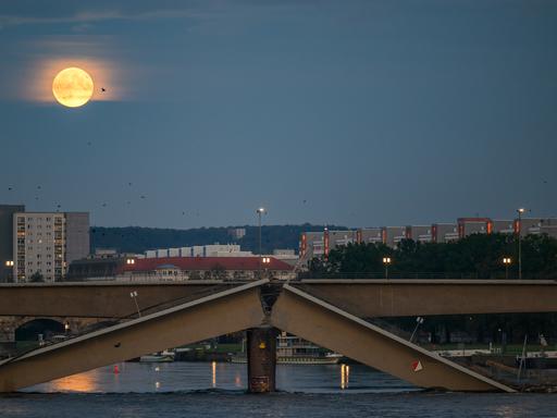 Der Vollmond ist am Abend am Himmel über der teileingestürzten Carolabrücke. Ein Strang der Brücke ist gebrochen, die Bruchstelle zeigt nach oben. Im Hintergrund sind Häuser.