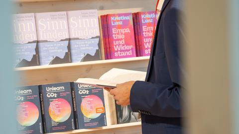 Eindrücke von der Frankfurter Buchmesse 2024. Im Anschnitt steht ein in einem Buch blätternder Mann vor einem Bücherregal mit Sachbüchern. 