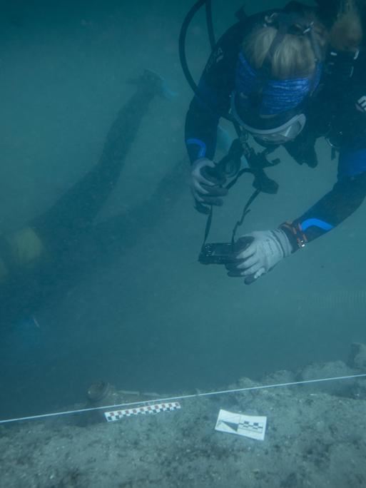 Ein Mensch in einem Tauchanzug fotografiert eine archäologische Stätte am Meeresgrund.