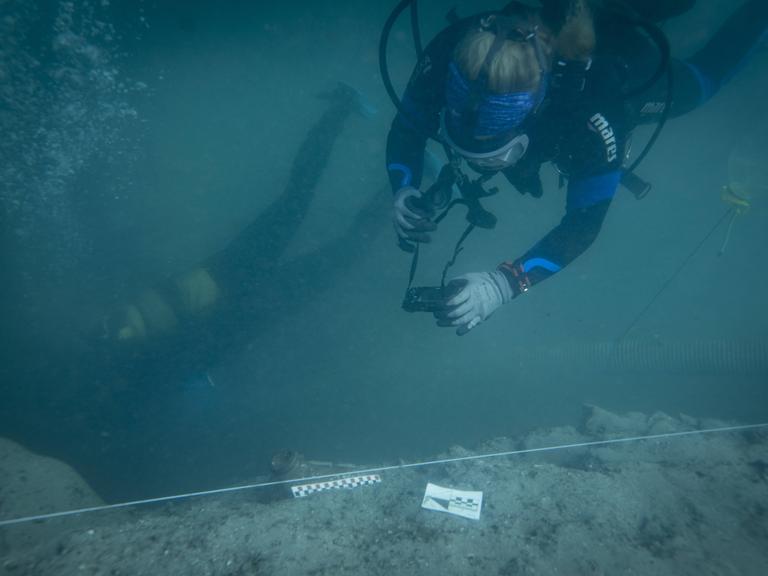 Ein Mensch in einem Tauchanzug fotografiert eine archäologische Stätte am Meeresgrund.