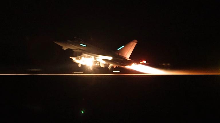 Es ist Nacht. Das gerade abhebende Flugzeug wird vom eigenen Jet-Strahl und von einem Scheinwerfer beleuchtet.