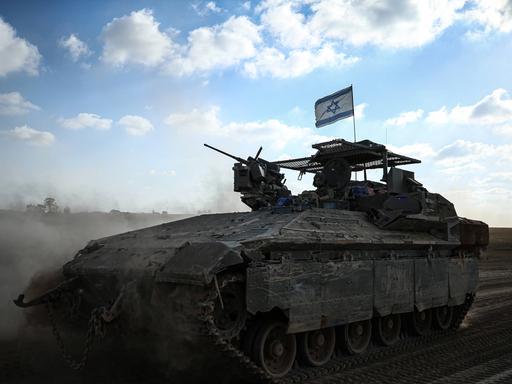 Ein israelisches Panzerfahrzeug mit Flagge wirbelt vor blauem Himmel Wüstensand auf.