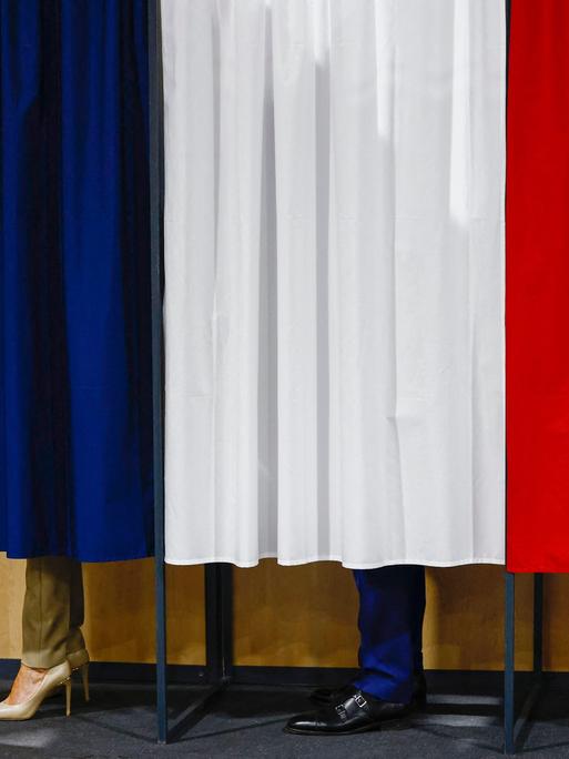 Unter einem Vorhang in den Farben der französischen Nationalflagge schauen die Beine und Schuhe von Emmanuel Macron und seiner Frau Brigitte hervor.