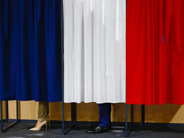 Unter einem Vorhang in den Farben der französischen Nationalflagge schauen die Beine und Schuhe von Emmanuel Macron und seiner Frau Brigitte hervor.