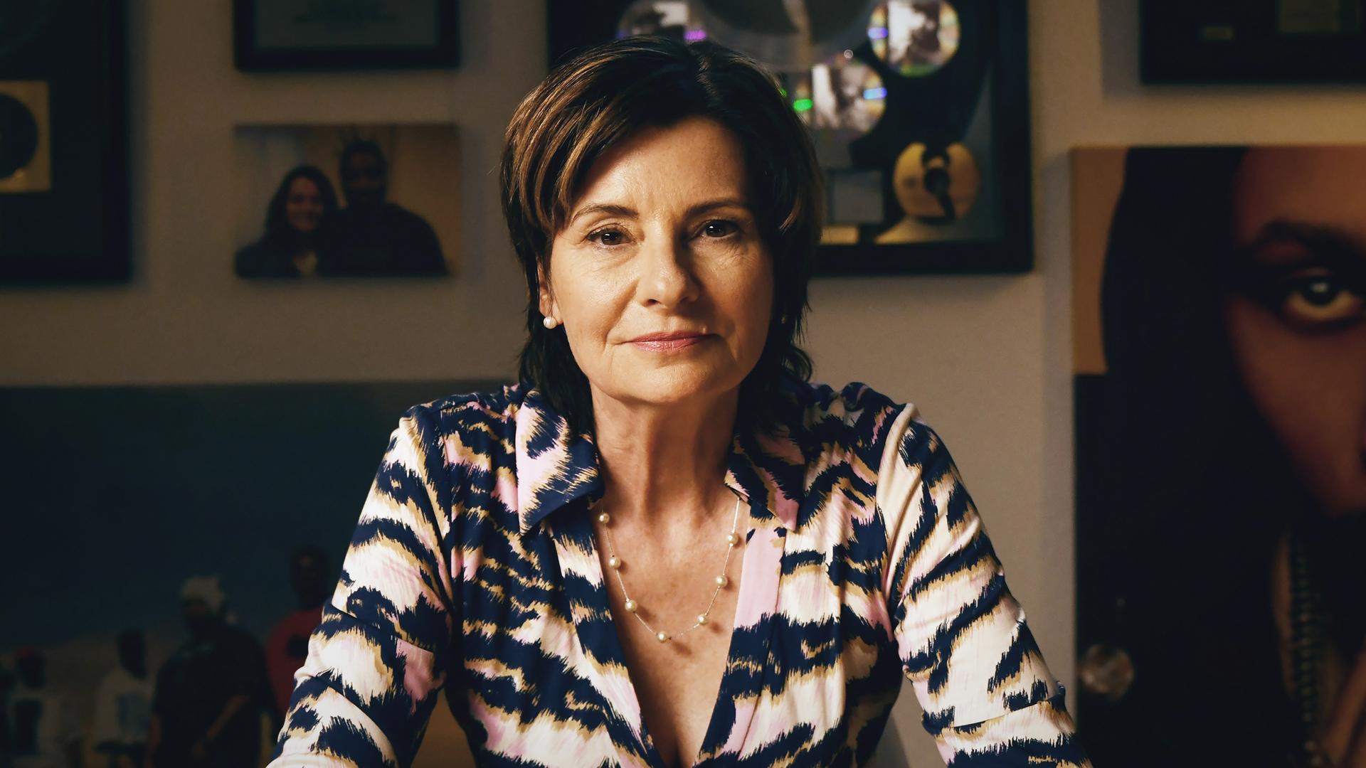 Eva Ries sitzt vor einer Wand mit gerahmten Bilder, trägt eine schwarz-weiße Bluse und schaut direkt in die Kamera. Eva Ries sitzt vor einer Wand mit gerahmten Bilder, trägt eine schwarz-weiße Bluse und schaut direkt in die Kamera.