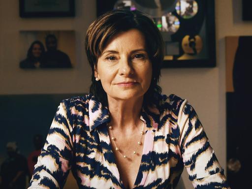 Eva Ries sitzt vor einer Wand mit gerahmten Bilder, trägt eine schwarz-weiße Bluse und schaut direkt in die Kamera. Eva Ries sitzt vor einer Wand mit gerahmten Bilder, trägt eine schwarz-weiße Bluse und schaut direkt in die Kamera.