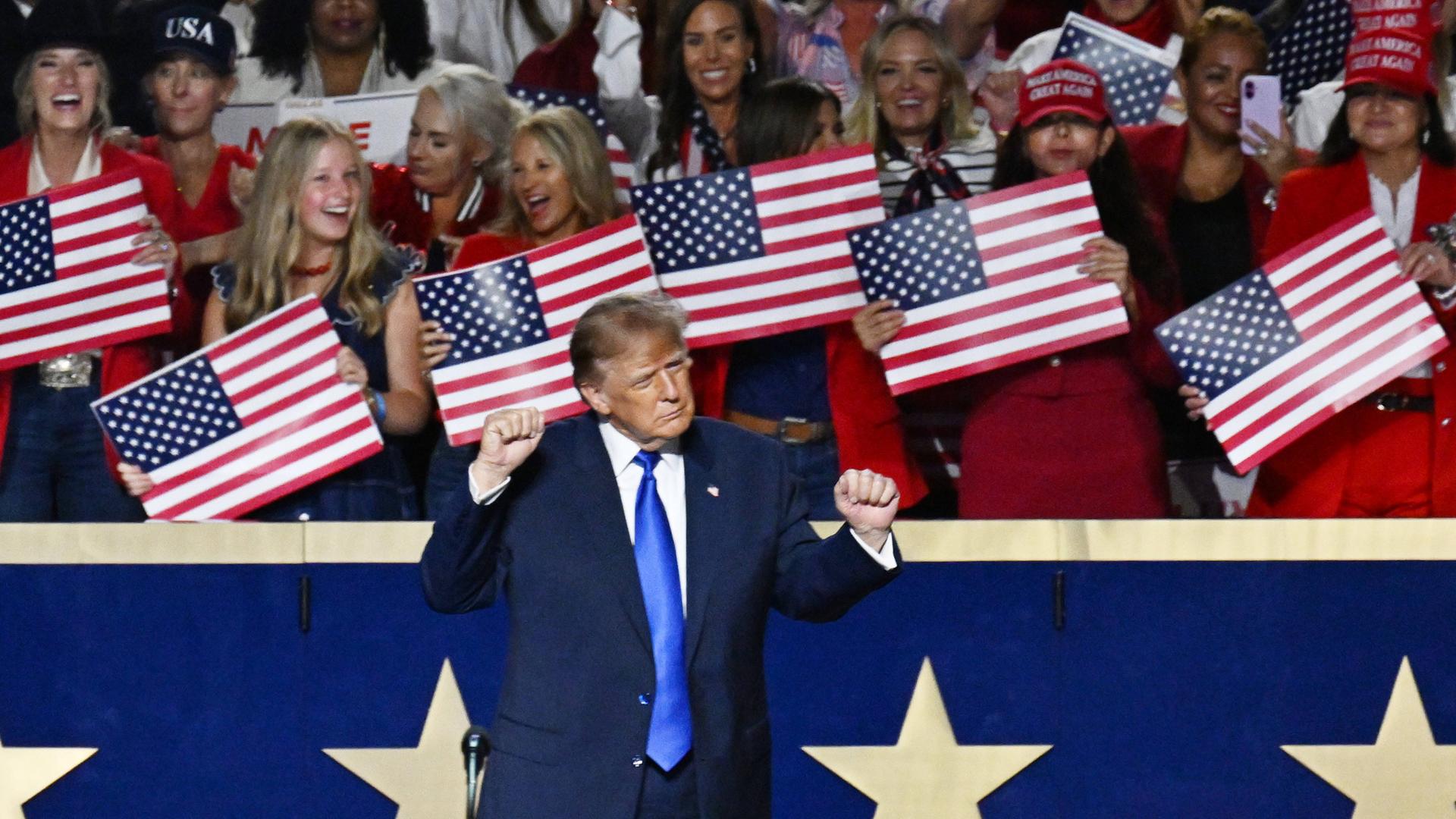 US-Präsident Donald Trump steht vor einer blauen Balustrade mit weißen Sternen und reckt beide Hände in die Höhe. Hinter ihm stehen Frauen, die kleine US-Flaggen in den Händen halten.