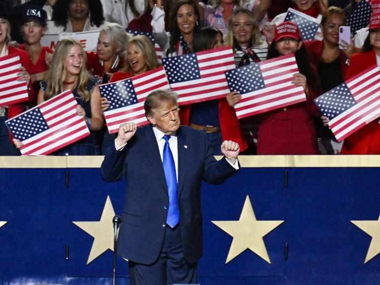 US-Präsident Donald Trump steht vor einer blauen Balustrade mit weißen Sternen und reckt beide Hände in die Höhe. Hinter ihm stehen Frauen, die kleine US-Flaggen in den Händen halten. US-Präsident Donald Trump steht vor einer blauen Balustrade mit weißen Sternen und reckt beide Hände in die Höhe. Hinter ihm stehen Frauen, die kleine US-Flaggen in den Händen halten.