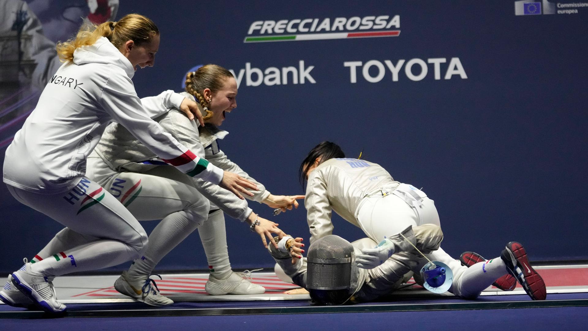 Das ungarische Frauen-Team jubelt: Ein Teammitglied liegt auf dem Boden, die anderen eilen zu ihr. Alle tragen Fecht- oder Sport-Kleidung. Im Vordergrund liegt ein Fecht-Visier. Das ungarische Frauen-Team jubelt: Ein Teammitglied liegt auf dem Boden, die anderen eilen zu ihr. Alle tragen Fecht- oder Sport-Kleidung. Im Vordergrund liegt ein Fecht-Visier.