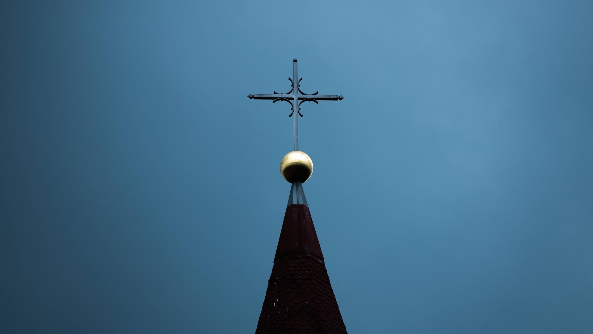 Die Spitze eines Kirchturms mit einer goldenen Kugel und einem dunklen Kreuz vor einem dunklen Himmel