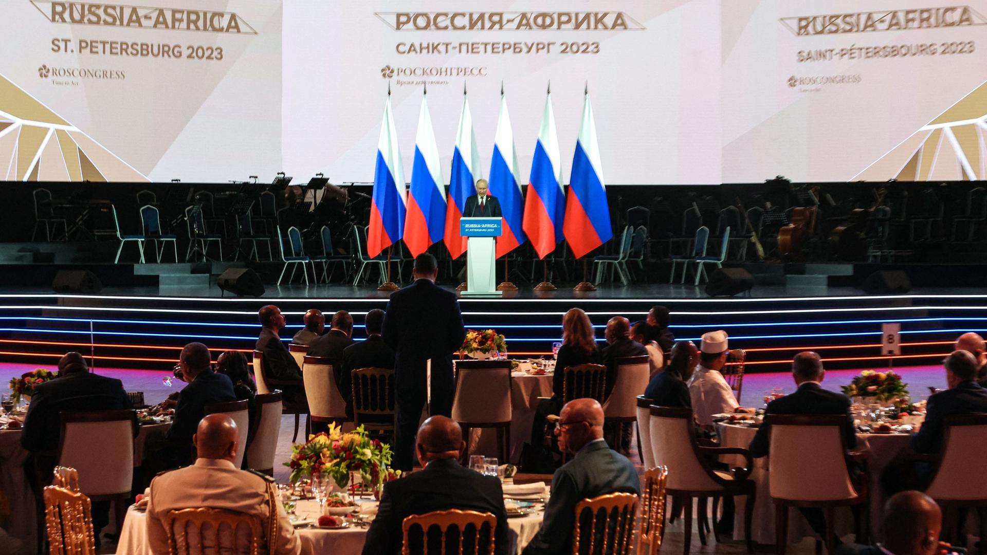 Präsident Putin steht beim Russland-Afrika-Gipfel in Sankt Petersburg auf der Bühne hinter einem Rednerpult. Hinter ihm mehrere russische Fahnen, vor ihm sitzen an runden Tischen Teilnehmer des Gipfels. Präsident Putin steht beim Russland-Afrika-Gipfel in Sankt Petersburg auf der Bühne hinter einem Rednerpult. Hinter ihm mehrere russische Fahnen, vor ihm sitzen an runden Tischen Teilnehmer des Gipfels.