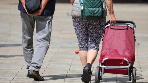Ein Seniorenpaar in luftiger Freizeitkleidung geht mit einem Einkaufstrolley durch die Leipziger Innenstadt. 
