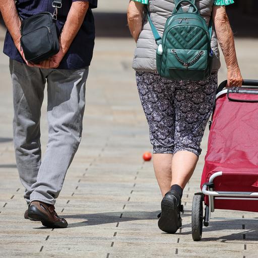 Ein Seniorenpaar in luftiger Freizeitkleidung geht mit einem Einkaufstrolley durch die Leipziger Innenstadt. 