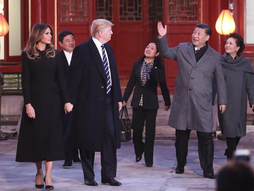 Donald Trump hält seine Frau Melania Trump an der Hand. Er blickt über seine Schulter zu Chinas Präsidenten X Jinping, der ihn verabschiedet.