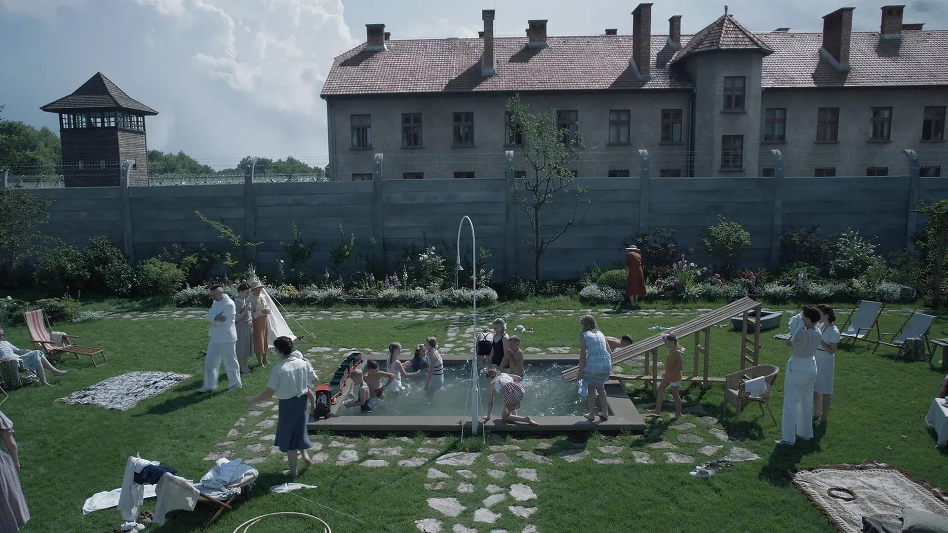 Filmszene aus "Zone of Interest". Zu sehen ist in einer Totale der Garten der Familie Höß mit Pflanzen und einem Pool, in dem Kinder fröhlich spielen. Im Hintergrund eine Mauer und ein Wachturm des sogenannten Stammlagers in Auschwitz.