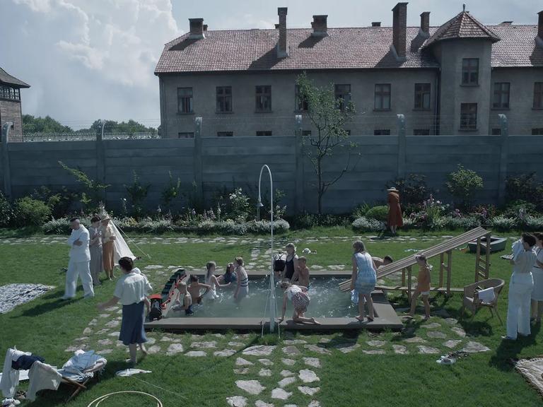 Filmszene aus "Zone of Interest". Zu sehen ist in einer Totale der Garten der Familie Höß mit Pflanzen und einem Pool, in dem Kinder fröhlich spielen. Im Hintergrund eine Mauer und ein Wachturm des sogenannten Stammlagers in Auschwitz.