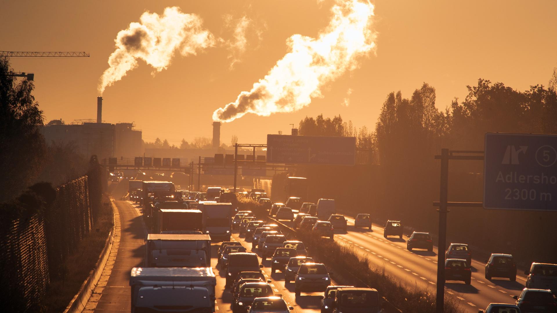 Das Bild zeigt eine Autobahn in den frühen Morgenstunden und große Schornsteine mit rauch im Hintergrund