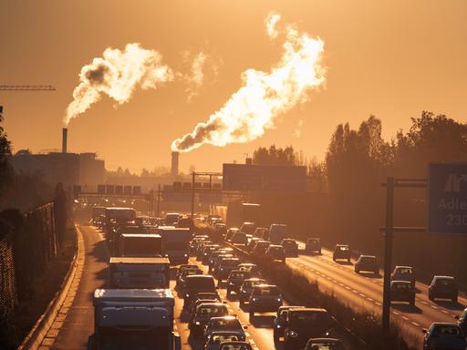 Das Bild zeigt eine Autobahn in den frühen Morgenstunden und große Schornsteine mit rauch im Hintergrund