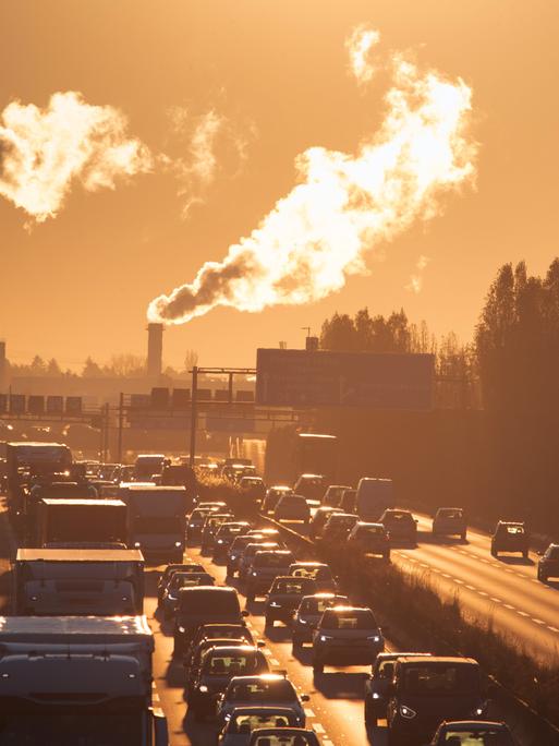 Das Bild zeigt eine Autobahn in den frühen Morgenstunden und große Schornsteine mit rauch im Hintergrund