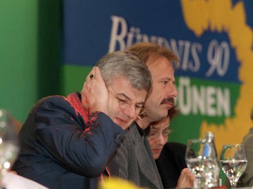 Joschka Fischer hält mit schmerzverzerrtem Gesicht seine Hand ans Ohr. Gesicht und Jackett sind mit roter Farbe beschmiert.