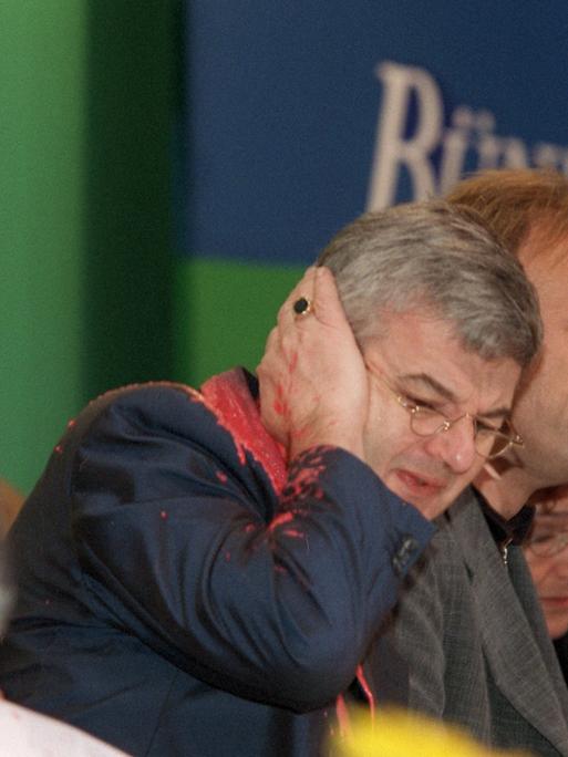 Joschka Fischer hält mit schmerzverzerrtem Gesicht seine Hand ans Ohr. Gesicht und Jackett sind mit roter Farbe beschmiert.