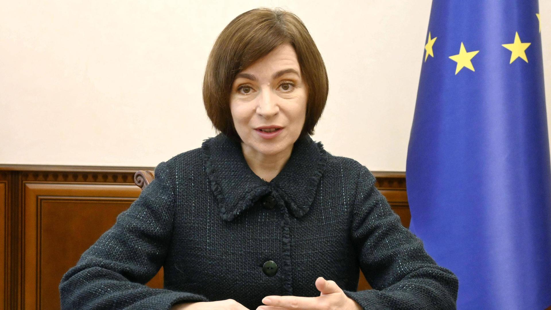 Maia Sandu, die Präsidentin Moldaus sitzt an einem Holztisch. Sie schaut in die Kamera. Ihre Hände liegen auf dem Tisch. Im Hintergrund sieht man angeschnitten eine EU-Flagge.