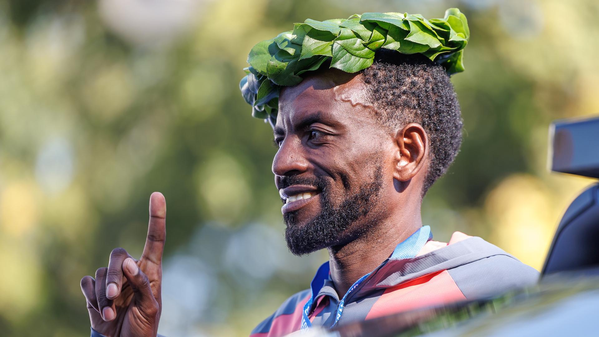 Siegerehrung beim Marathon in Berlin: Man sieht Guye Adola von der Seite- Er trägt einen Lorbeerkranz auf dem Kopf, lächelt und hebt den Zeigefinger der rechten Hand. 