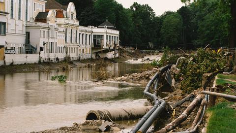 Links ist ein Gebäude zu sehen, das dicht an einem Fluss gebaut ist. Im Vordergrund der braun gefärbte Fluss und frei liegende Rohre am Ufer, das dem Gebäude gegenüber liegt.