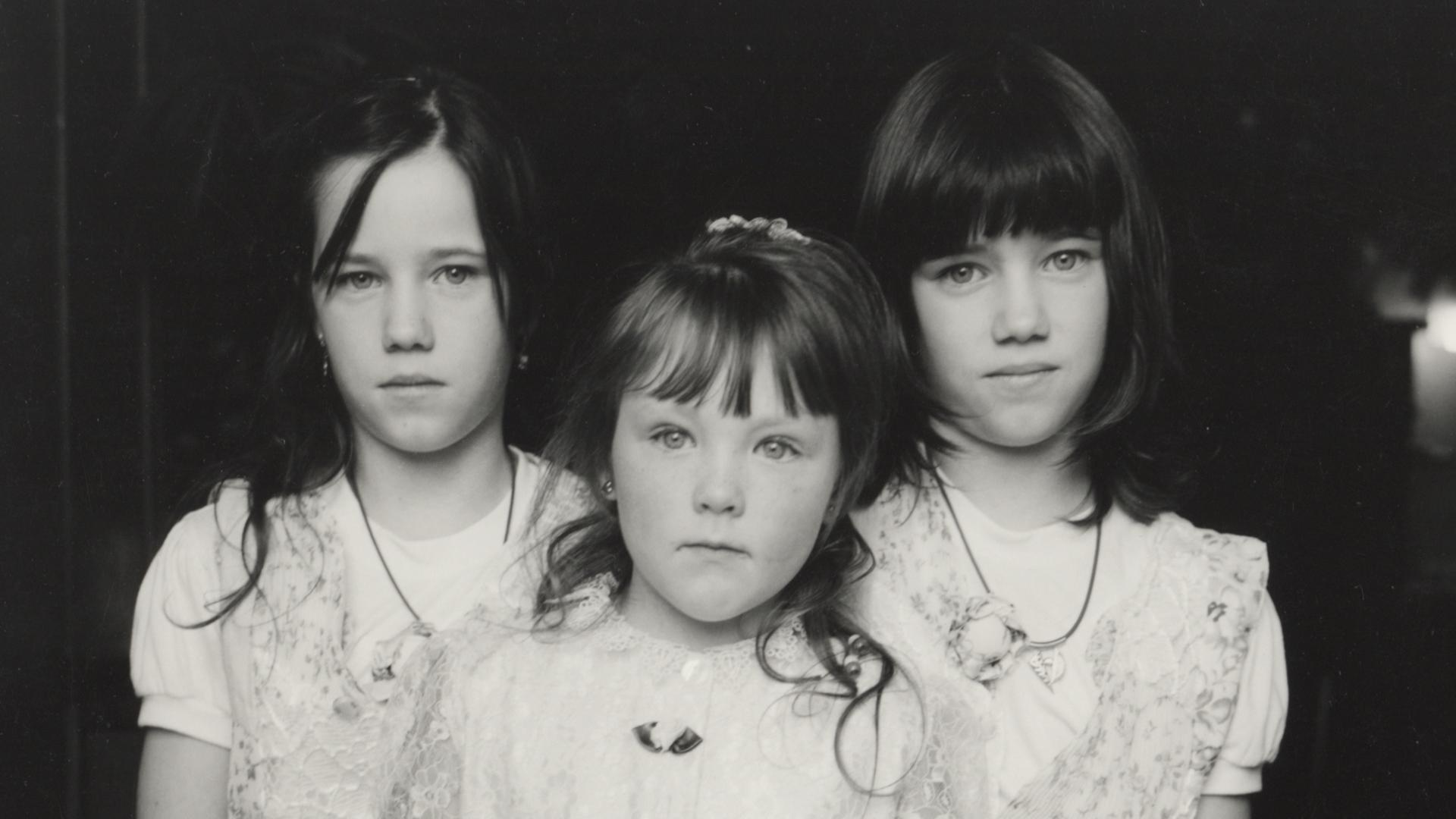 Schwarz-weiß Foto von drei kleinen Schwestern in weißen Kleidern, die skeptisch in die Kamera gucken.