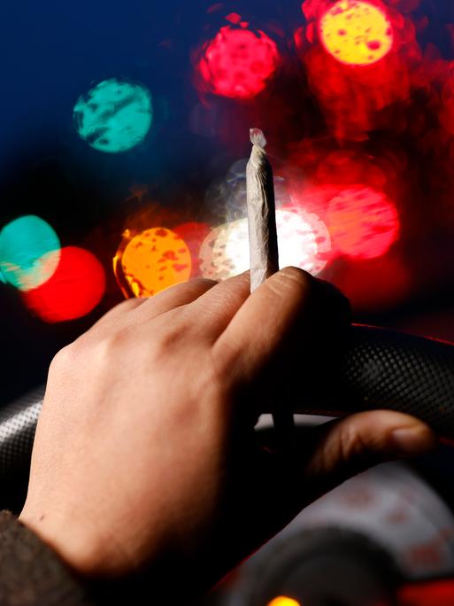 Eine Hand mit einem Joint hält ein Lenkrad, im Vordergrund sind unscharf die Lichter des Straßenverkehrs erkennbar. 