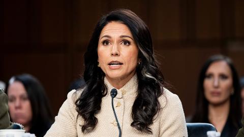 Gabbard sitzt an einem Tisch und spricht in ein Mikrofon. Sie trägt eine helle Jacke.