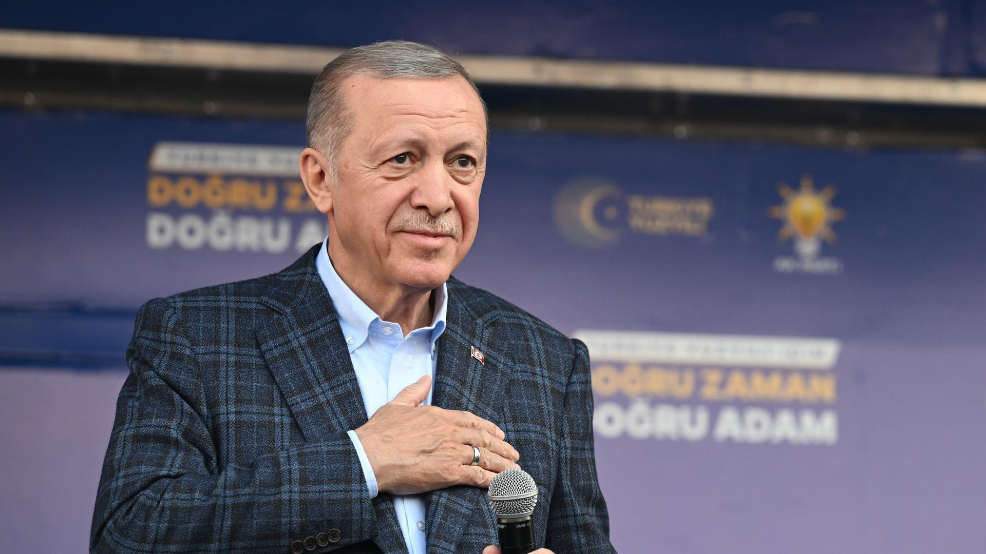 Porträtbild des türkischen Präsidenten Recep Tayyip Erdogan. Er steht mit einem Mikrofon in der Hand auf einer Bühne und hat seine rechte Hand auf seine Herzgegend gelegt. 