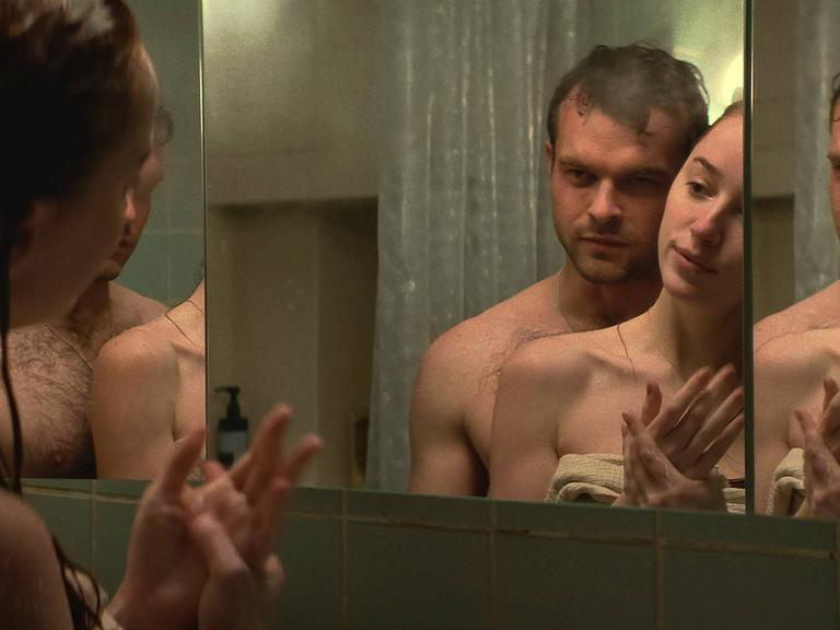 Eine Mann und eine Frau stehen leicht bekleidet vor einem Spiegel.