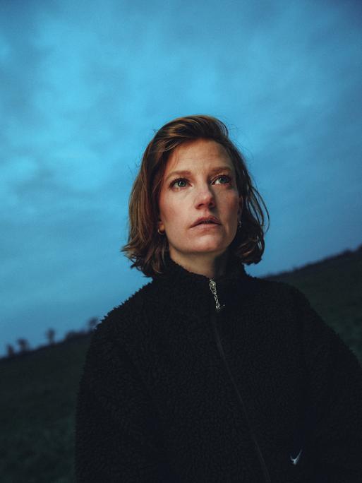 Die Sängerin Wilhelmine steht mit schwarzem Pullover auf einem Feld bei einbrechender Dämmerung und schaut fragend Richtung Kammera. 