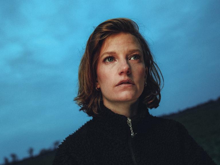 Die Sängerin Wilhelmine steht mit schwarzem Pullover auf einem Feld bei einbrechender Dämmerung und schaut fragend Richtung Kammera. 